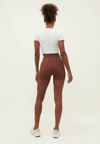 Ruskeat, korkeavyötäröiset shortsit joustavasta materiaalista, yhdistetty istuvaan valkoiseen crop-topiin, käytetty vaalean väristen urheilukenkien kanssa.