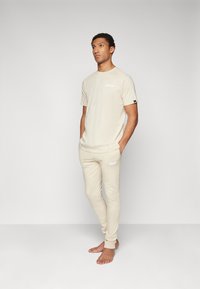 T-shirt en coton beige et joggings assortis. Coupe ajustée avec logo sur le t-shirt et le pantalon. Texture lisse, col rond et poignets élastiques.