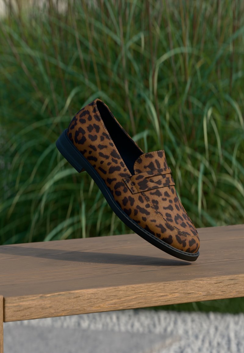 Mocassin à imprimé léopard marron et noir posé sur le bord d'un banc en bois, avec des hautes herbes vertes en arrière-plan.