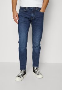 Blå denimjeans med slim fit, som har fem fickor och kontrasterande orange sömmar, tillsammans med svarta högskaftade sneakers.
