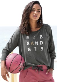 Dunkelgraues Langarmshirt mit weißer und goldener Schrift, kombiniert mit pinken Sporthosen. Hält einen pinken Basketball und steht im Freien.