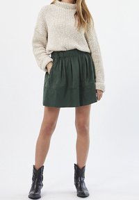 Jupe en daim verte avec une taille, des plis et des poches ; associée à un pull en tricot chunky beige clair et des bottes de cowboy noires.