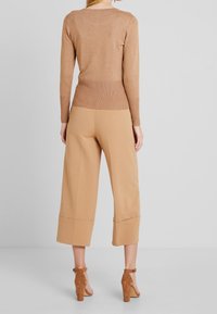 Frau steht mit dem Rücken zur Kamera, trägt ein eng anliegendes, braunes Langarmshirt, beige, weit geschnittene Cropped-Hosen und braune High Heels.