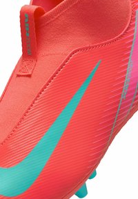 Botas de fútbol rojas con parte superior de punto, que presentan un gran swoosh en aqua, superficie texturizada y una lengüeta etiquetada como "AIR ZOOM". Clavos suaves en la suela.