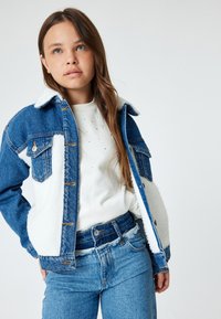 Chaqueta de mezclilla con forro de sherpa blanco, tonos azules, dos bolsillos en el pecho y cierres de botones. Llevada sobre un suéter blanco y jeans azules.