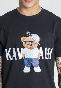 Černé tričko s potiskem postavičky medvěda v brýlích a kšiltovce, oblečeného v džínách a bílých botách, spolu s nápisem "KAVACH."