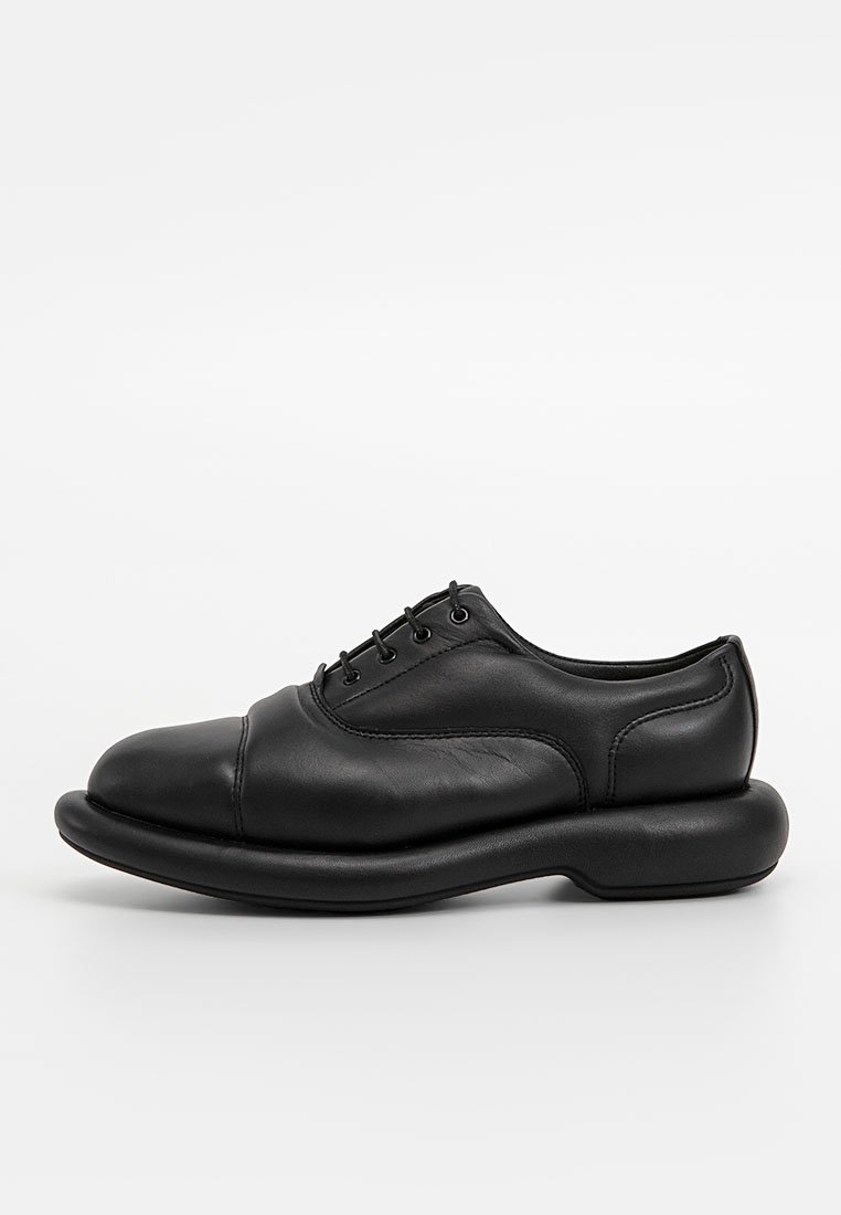 Chaussure en cuir noir avec un bout rond, un laçage et une semelle épaisse et arrondie au toucher lisse.