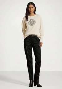 Γυναίκα φορώντας μπεζ φούτερ με μαύρο λογότυπο, μαύρα cargo παντελόνια με τσέπες, μαύρες γόβες με τακούνι στον αστράγαλο, στέκεται μπροστά σε λευκό φόντο.