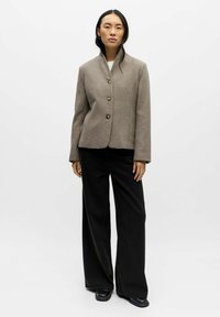 Bruine jas van een wolmix met een hoge kraag, drie knopen en een getailleerde pasvorm, gecombineerd met zwart wijduitlopende broeken en zwarte schoenen.