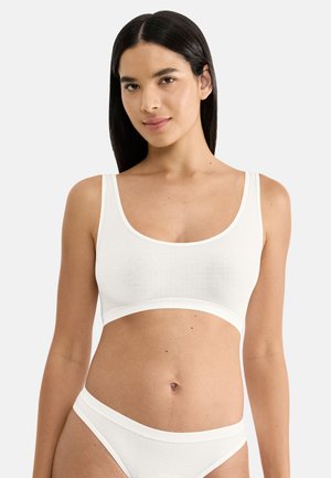 Frau mit langen dunklen Haaren, die ein weißes nahtloses Crop-Top und passende hochgeschnittene Unterwäsche trägt, steht vor einem schlichten hellen Hintergrund.