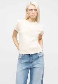 Νεαρή γυναίκα με ίσια ξανθά μαλλιά, φορώντας απλό εκρού κοντομάνικο μπλουζάκι και ανοιχτό μπλε τζιν, με τα χέρια στις τσέπες.