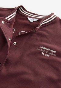 Burgundi värvi polo särk, millel on kraega kaelus, valged triibud ja tikitud tekst "Autentne osakond. Chicago kollektsioon asutatud 1995."