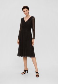 Zwarte jurk met lange, doorschijnende mouwen, een V-hals en een geraffineerde taille. Vloeiende rok met een geruit textuur. Gepaard met zwarte hoge hakken sandalen.