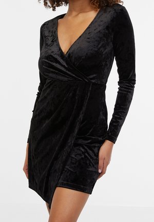 Femme portant une robe portefeuille en velours noir à manches longues, avec un profond décolleté en V et un ourlet court, sur un fond uni.