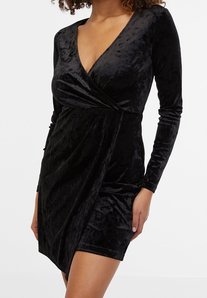 Femme portant une robe portefeuille en velours noir à manches longues, avec un profond décolleté en V et un ourlet court, sur un fond uni.