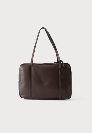 Dunkelbraune Lederhandtasche mit zwei langen Griffen und einem kleinen angebrachten rechteckigen Etikett auf einem schlichten hellen Hintergrund.