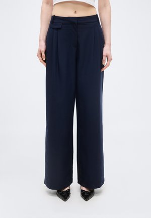 Kobieta w granatowych, szerokich spodniach z wysokim stanem i plisami z przodu oraz czarnych szpilkach z zakrzywionym noskiem, stojąca na jednolitym tle.