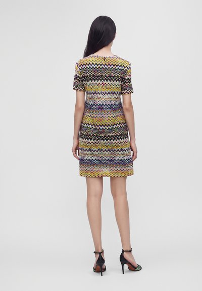 Femme aux longs cheveux foncés portant une robe à manches courtes à motif zigzag multicolore et des sandales noires à talons hauts, debout de dos.