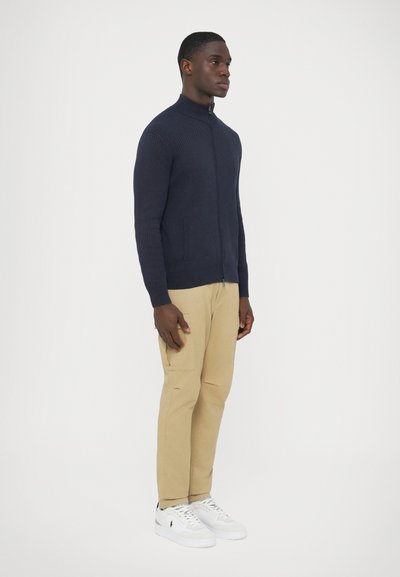 Sweater azul-marinho com fecho de correr, textura canelada, combinado com calças cáqui claras e ténis brancos com detalhes pretos.