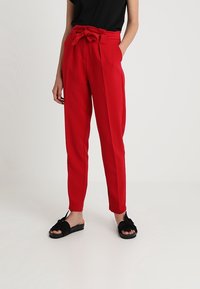 Red high-waisted trousers with a fabric belt, featuring side pockets and a tailored fit. Worn with a black top and black flat sandals.