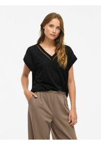 Frau mit langen, welligen Haaren, die ein schwarzes V-Ausschnitt-Top mit Spitzenakzenten und lockere braune Hosen trägt, steht mit einer Hand in der Tasche.