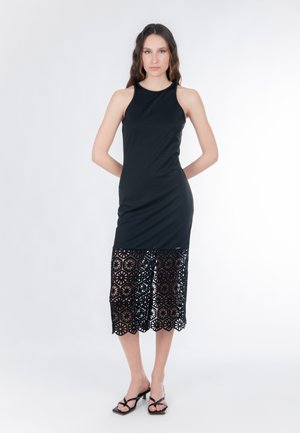 Vrouw staat met handen achter haar rug, draagt een mouwloze zwarte jurk met een kantpatroon over de rok en zwarte sandalen met bandjes.