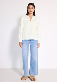 Blusa bianca a maniche lunghe con scollatura a V e dettagli in pizzo sulle spalle, abbinata a jeans a gamba larga blu chiaro e sandali beige.