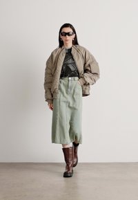 Pufferjakke i taupe, svart tettsittende skjorte, lys grønn midi-skjørt, og brune knelang støvler med tykke såler, modell som står på en ensfarget bakgrunn.