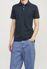 Tmavě modré polo triko s klasickým límečkem, třemi knoflíky a malým červeným logem na hrudi. Spárováno se světle modrými džíny.