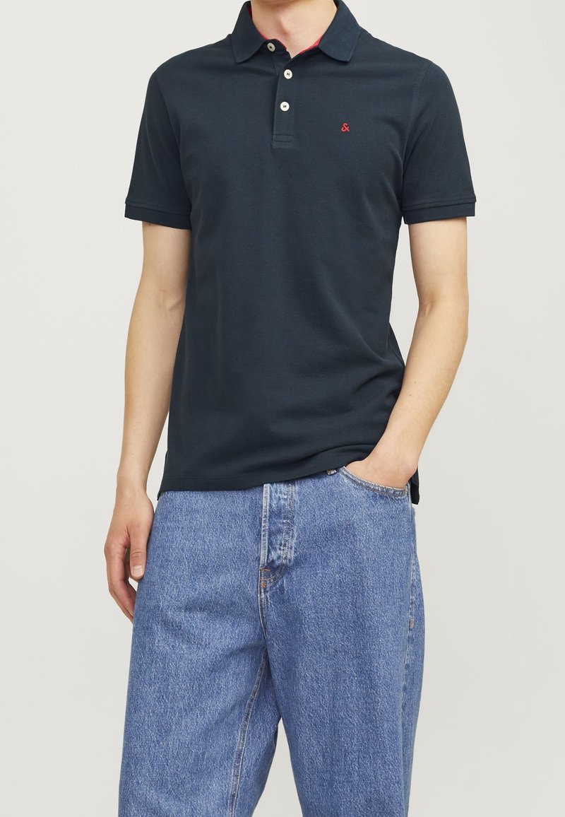 Tmavě modré polo triko s klasickým límečkem, třemi knoflíky a malým červeným logem na hrudi. Spárováno se světle modrými džíny.