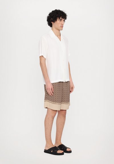 Camicia bianca a maniche corte, pantaloni corti marroni a fantasia con rifiniture beige e sandali neri. Modello in piedi con un'espressione neutra su uno sfondo bianco.