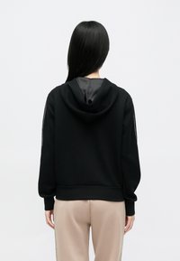 Μαύρο φούτερ με κουκούλα, με λεία υφή, ριμπ μανσέτες και τελείωμα. Διαθέτει φερμουάρ στο πίσω μέρος και άνετη γραμμή. Φορεμένο με ανοιχτόχρωμο παντελόνι.