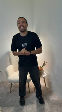Homme souriant et joignant les mains, portant un t-shirt noir et un pantalon, debout dans une pièce avec moquette, une chaise blanche et une petite table tenant un vase et des livres.