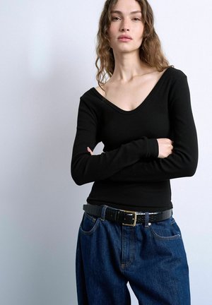 Young woman with wavy hair, wearing a fitted black long-sleeve top and loose blue jeans with a black belt, standing against a plain background.