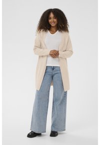 Moteris su garbanotais plaukais, apsirengusi smėlio spalvos švarku, baltu marškinėliu, plačiomis šviesiai mėlynomis džinsomis ir juodais bateliais, stovi su susikirtusiomis rankomis.