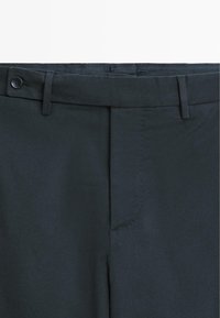 Tmavě zelené kalhoty vyrobené z strukturované tkaniny. Mají zapínání na knoflík, poutka na opasek a ostré záhyby vpředu. Hladký povrch.