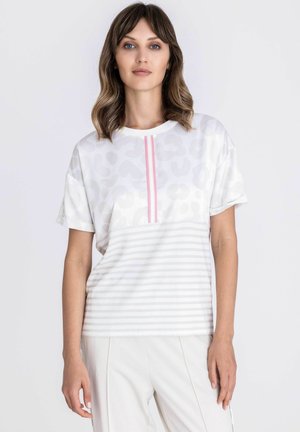 Junge Frau mit welligem braunem Haar trägt ein weißes kurzärmeliges Oberteil mit grauen Leoparden- und Streifenmustern und einem pinken vertikalen Streifen.