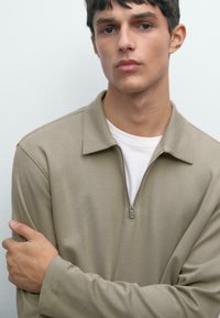 Ung man med kort mörkt hår, iförd en beige zip-up långärmad skjorta över en vit T-shirt, som står framför en enkel ljus bakgrund.