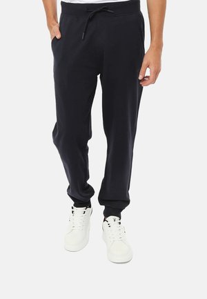 Uomo che indossa pantaloni jogger neri con coulisse in vita e sneaker bianche, in piedi con una mano in tasca su sfondo bianco.