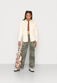 Camisa creme de botão sobre um top cropped castanho, combinada com calças cargo camufladas verdes. Sapatos pretos e um saco tote com padrão de várias cores.