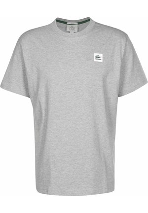T-shirt gris clair à manches courtes avec col rond, petit écusson logo Lacoste sur la poitrine gauche et détail vert à l’intérieur du col.