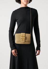 Guldvävd handväska med rektangulär form, med en stor spänne och ett kedjehandtag, hålls av en person som bär en svart långärmad klänning.