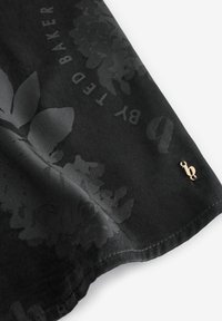 Musta huivi, jossa on kukkakuosi ja logo hienovaraisissa sävyissä. Reunat on hienosti ommeltu, ja yhdessä kulmassa on kultasävyinen yksityiskohta.