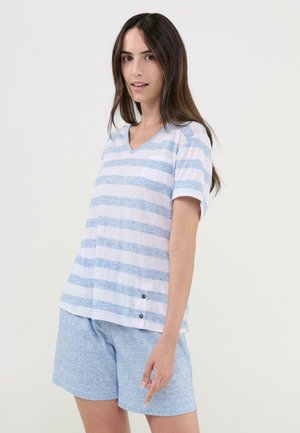 Mujer con camiseta de manga corta a rayas blancas y azul claro y pantalones cortos azul claro, de pie contra un fondo liso claro.