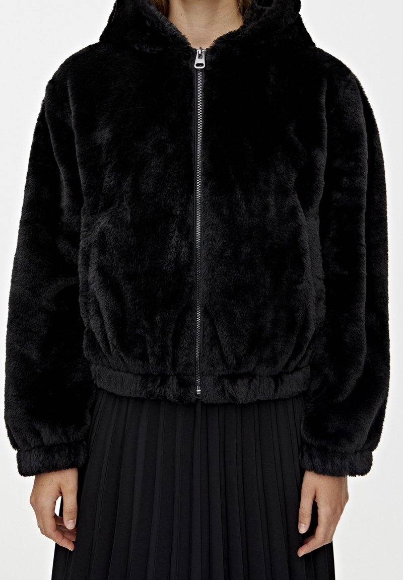 Black faux fur hoodie with a zip closure, elasticated waistband, and long sleeves, paired with a black pleated skirt. Soft texture and cozy design.