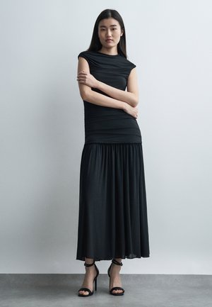 Jeune femme debout contre un mur uni, portant une robe noire sans manches et des sandales noires à talons hauts, les bras croisés.