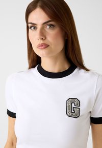 Biała koszula z krótkim rękawem z czarnym prążkowanym kołnierzykiem i mankietami. Na piersi znajduje się teksturowane, czarno-srebrne ozdobne logo "G".