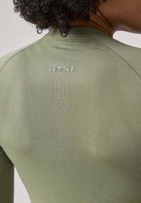 Grünes Sportoberteil aus strukturiertem, atmungsaktiven Stoff. Verfügt über ein figurbetontes Design, mit dem 'SNOCKS'-Logo in hellgrün im oberen Rückenbereich.