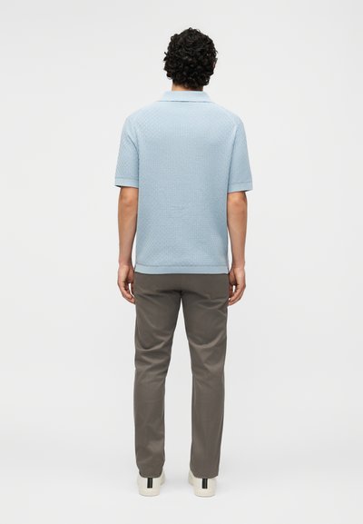 Άνδρας με σγουρά σκουρόχρωμα μαλλιά, γυρισμένος προς τα πίσω, φοράει ανοιχτό μπλε πλεκτό μπλουζάκι με κοντά μανίκια, γκρι παντελόνι και λευκά αθλητικά παπούτσια με μαύρες ρίγες.