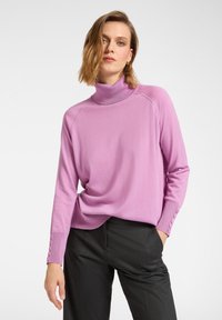 Różowy sweter z golfem i długimi rękawami, ozdobiony guzikami na mankietach, o luźnym kroju. Noszony z ciemnoszarymi spodniami.